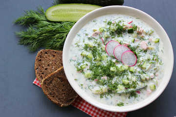 Okroshka is a traditional Russian summer cold soup in a ceramic bowl. Ingredients potatoes, radishes, cucumbers, dill, eggs, kefir (yogurt), sausage. Summer yogurt cold soup on a wooden table. Selecti
