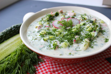 Okroshka is a traditional Russian summer cold soup in a ceramic bowl. Ingredients potatoes, radishes, cucumbers, dill, eggs, kefir (yogurt), sausage. Summer yogurt cold soup on a wooden table. Selecti