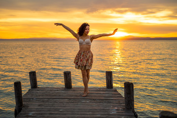 Fototapeta premium Happy woman enjoying the summery sunset at the lake on her vacation. The woman in bra and a short summer skirt laughs and puts her arms in the direction of the sky