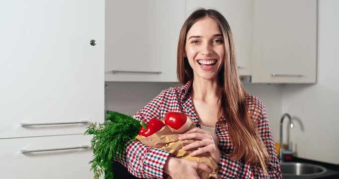 Portrait of young pretty woman holding paper pack of fresh vegetables before kitchen background