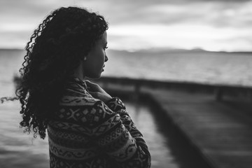 Thoughtful evening mood with a young African American woman standing on the promenade by the lake and looking towards the water and the setting sun. Photo in black and white