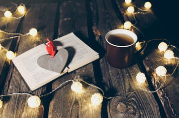 Holidays mood photo. Christmas lights and hot tea mug. Book for cosy evening. Sweet gingerbread and wooden heart on tray. Perfect winter flat lay. Hygge concept.