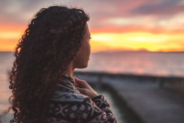 Afro American woman standing on the promenade by the lake and looking towards the water and the setting sun. Burning sky. Thoughfull moment