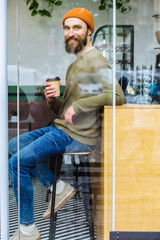 Vertical portrait of positive handsome hipster man wears stylish clothes, drinks coffee at cafeteria, looks at camera through window glass, has serious expression. Sporty man relaxes working day.