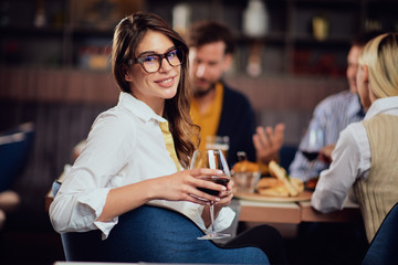 Portrait of gorgeous brunette looking at camera and holding glass with red wine while sitting at restaurat. In background friends eating diner.