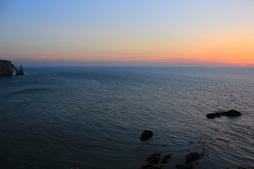 The Etretat cliffs (Normandy) in France. They are only present through the low key profiles of the Natural Arch and of the  Needle. The August sun sets on the dark blue waters of the English Channel.