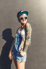 Freestyle. Alternative girl in cap ans sunglasses standing isolated on grey on the city street...