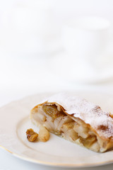 Apple strudel served on a white porcelain plate. Coffee cups in bokeh, white table, high resolution