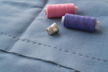 Fototapeta na wymiar Cut part of skirt with a sewn tuck, thimble and two spools of thread. Two spools of pink and purple threads and tailor tool are lying on the blue basting fabric