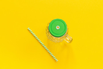 Empty glass jar in form of pineapple with green lid and straw for fruit or vegetable smoothies, cocktails and other beverages on yellow background. Top view Copy space Template