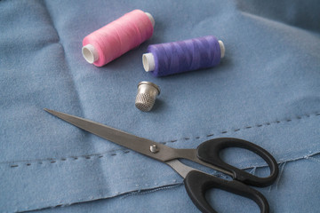 Top of view of the cut part of skirt with a sewn tuck, scissors, thimble and two spools of thread. Two spools of pink and purple threads, metal cap and scissors are lying on the blue fabric