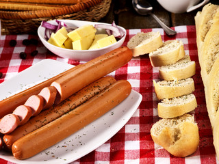 Sausage portion with bread baguette on table setting on checkered tablecloth and cup in farm style. Cozy, homely atmosphere.