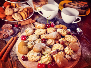 Oatmeal cookies and sand chocolate cake with cherry berry on cutting board on wooden table in rustic style. Top view of still life served with cups and dessert spoons. Sun flare morning sunrays.