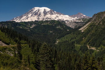 A snow capped mountain, Mount Rainier, at spring time with a forest of lush green pine trees in the foreground and road sweeping across the picture, nobody in the image