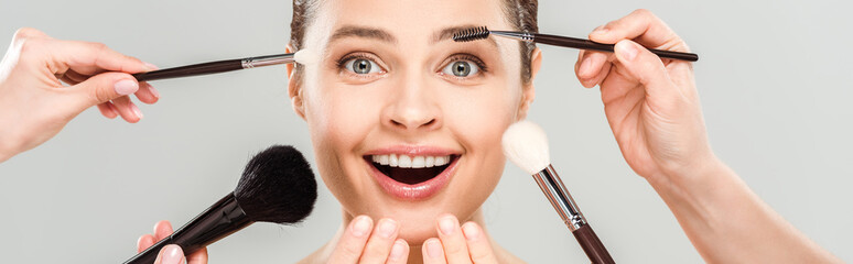 panoramic shot of makeup artists holding cosmetic brushes near excited woman isolated on grey