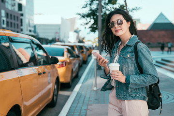 confident businesswoman using application of taxi service app on smart phone holding coffee to go. young asian office lady with backpack calling cab on cellphone standing on street smiling outdoor.