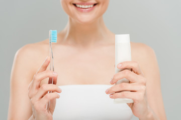 cropped view of woman smiling while holding toothbrush and tube with toothpaste isolated on grey