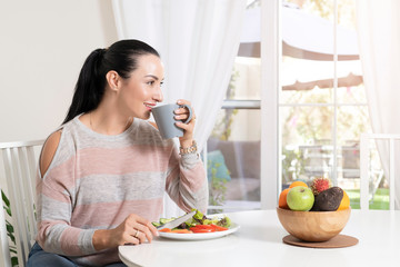 Obraz na płótnie Canvas Beautiful brunette woman sitting at dining table eating a healthy salad and drinking coffee or tea with a bowl of fruits on table with garden in the background