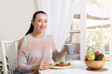 Beautiful brunette woman sitting at dining table eating a healthy salad and drinking coffee or tea with a bowl of fruits on table with garden in the background