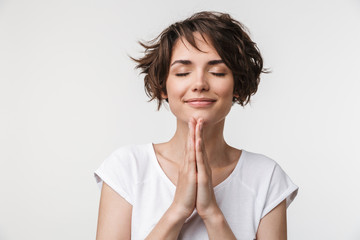 Portrait of joyous woman with short brown hair in basic t-shirt keeping palms together and praying - obrazy, fototapety, plakaty
