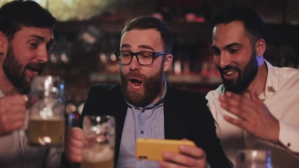 Three old friends watching video on smartphone and drinking draft beer in the sport bar. Friends laughing and discuss the news in beer pub.