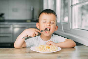 little boy eats pasta in the form of a spiral in the afternoon in the kitchen on their own