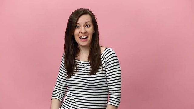 Smiling brunette young woman in striped shirt pointing in different directions, looking at camera with charming smile isolated over pastel pink background in studio. People emotions, lifestyle concept