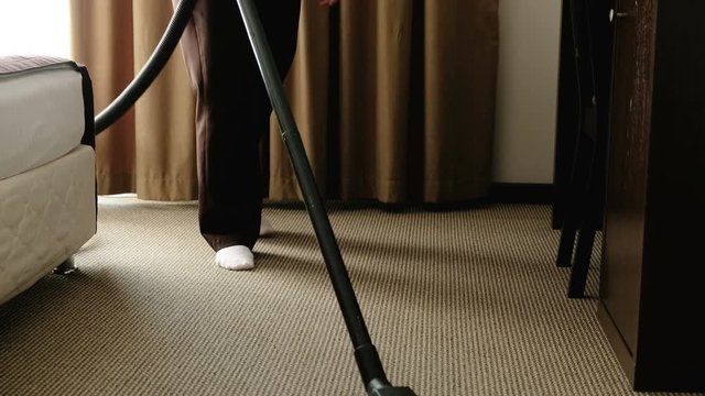 An unidentified middle-aged woman vacuums a carpet in a hotel room. The concept of high quality service and cleaning in the hotel room