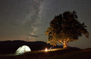 Female camper resting at summer night camping in the mountains beside campfire and tourist tent. Backpacker sitting on chair under big old tree and beautiful night sky full of stars and Milky way.