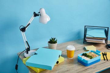 lunch box with healthy food and coffee in paper cup at workplace with folders and papers on wooden table on blue background