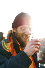 Handsome young man fisherman wearing coat and hat at the seashore drinking hot tea.