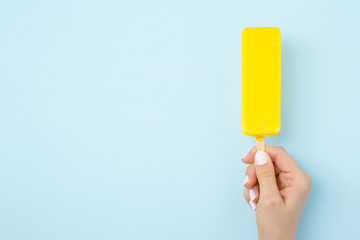 Young woman hand holding bright yellow ice cream with lemon glaze on light pastel blue background. Empty place for text, quote or sayings. Close up.  - obrazy, fototapety, plakaty