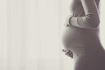 Pregnant thoughtful woman standing at window curtains in room. Single mother. Suffering from life...