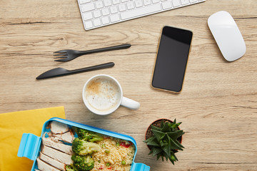 Top view of lunch box with delicious healthy food, hot beverage and digital devices on wooden table