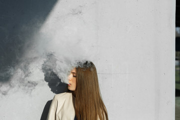Vape teenager. Young cute girl in  casual clothes smokes an electronic cigarette near the wall outdoors in summer day. Bad habit that is harmful to health. Vaping activity. Close up.