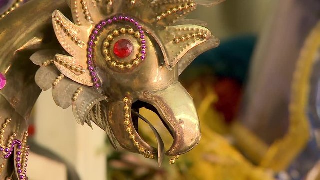Extreme close-up, low angle still shot of a decorated golden antique head of a tinsmith made rooster with red beads on its eye, surrounded by matching gold and purple beads, Oruro workshop, 