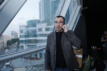 businessman is talking on his mobile phone with emotion of surprise on his face in the office opposite the panoramic window through which the street with buildings and cars is visible.  Copy space.