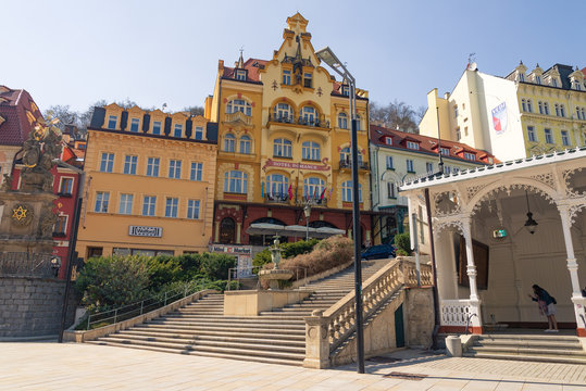 Outdoor sunny view of Holy trinity column, fountain and stairway beside Market colonnade, and background of Beautiful hotel's facade and colorful buildings in Karlovy Vary, Czech Republic..   - Powered by Adobe