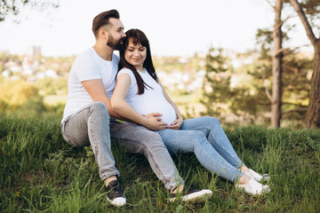 Naklejka na ściany i meble Happy young couple expecting baby, pregnant woman with husband touching belly, sitting on green grass