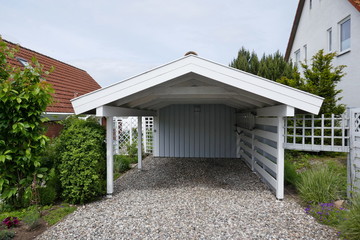 Wooden carport with pitched roof, white with open driveway on pebble floor next to a house....