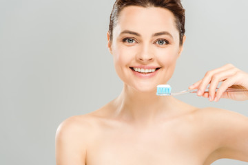 cheerful naked woman smiling while holding toothbrush with toothpaste isolated on grey