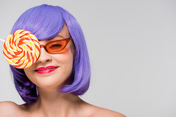 smiling girl in purple wig and sunglasses holding sweet lollipop, isolated on grey