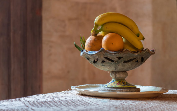 Un frutero antiguo cargado con pl&aacute;tanos y naranjas sobre una mesa