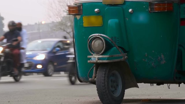 Motor-rickshaw of green color stands on the edge of the road, waits for clients, against the background of moving transport in Delhi, India