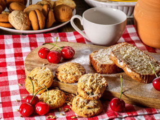Oatmeal cookies and cherry on kitchen cutting board gingham checkered cotton fabric on table in village style for picnic with plate of macaron background. Milk jug in background.