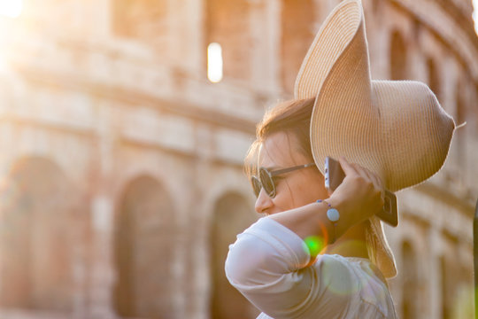 Beautiful young woman in fashion dress alone in front of colosseum in Rome at sunset. Taking off large hat.