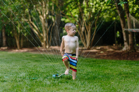 Cute young boy in swim trunks running through a water sprinkler
