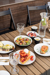 Stunning breakfast or lunch in open air in a cozy summer cafe. Poached eggs with Dutch sauce and avacado, coffee, bottle of water with mint, granola with fruit, waffles with ice cream, salad and chia 