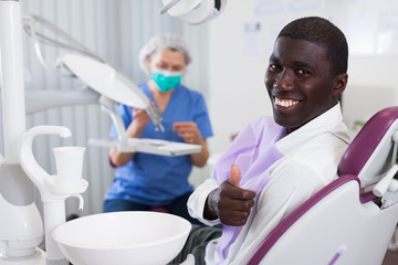 Happy african american man sitting in dental chair after teeth cure giving thumb up