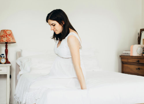 Thoughtful expectant woman sits on bed in bright bedroom